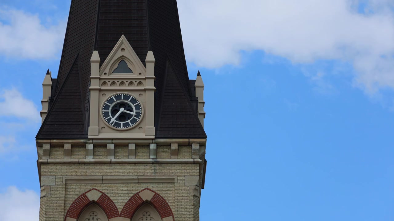 En stillbild från en timelapse-video som visar ett gammalt arkitektoniskt kyrkklocktorn. Klockans visare rör sig medan vita moln snabbt passerar över en blå himmel i bakgrunden under dagtid.
