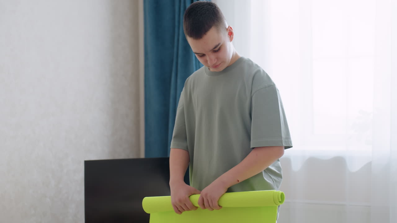 Teen boy folds bright green fitness mat inside cozy living room with television beside him and soft light filtering through curtains, showing calm focus and routine