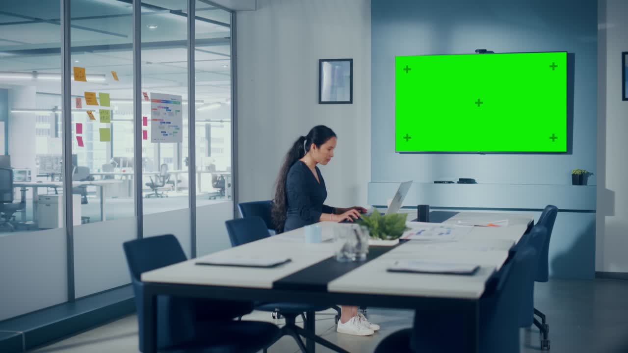 Office Conference Meeting Room: Beautiful Asian Specialist Working on Laptop Computer interacts with Green Screen Choma Key Wall TV. Businessperson Working in Growing e-Commerce Startup. Wide Shot