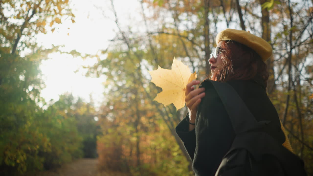 숲길을 고 있는 여인, 노란색 베레트와 선글라스를 입은 가방을 조정하고, 따뜻한 빛이 그녀를 둘러싸고 반이는 효과를 일으키며, 가을의 분위기와 평화로운 자연 풍경을 향상시니다.