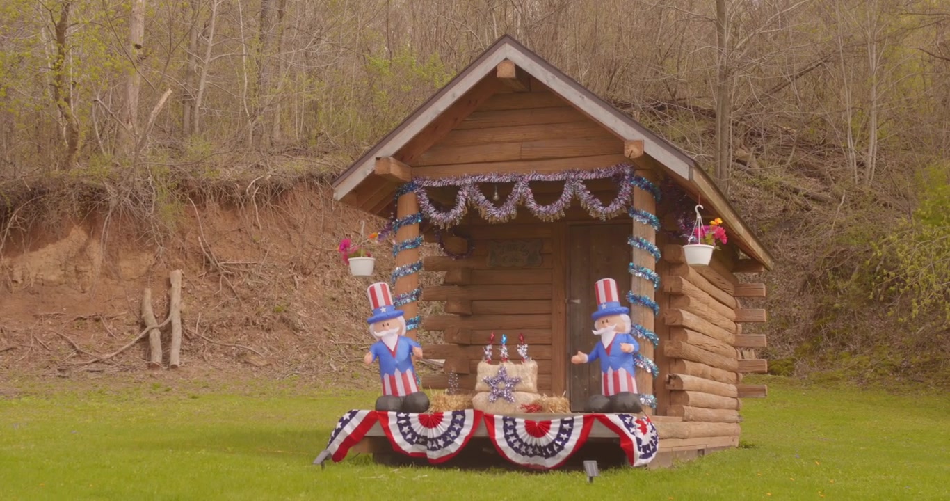 Small wooden log cabin on green grass decorated with red, white, and blue bunting with inflatables and a patriotic display terrace in a quiet woodland setting.