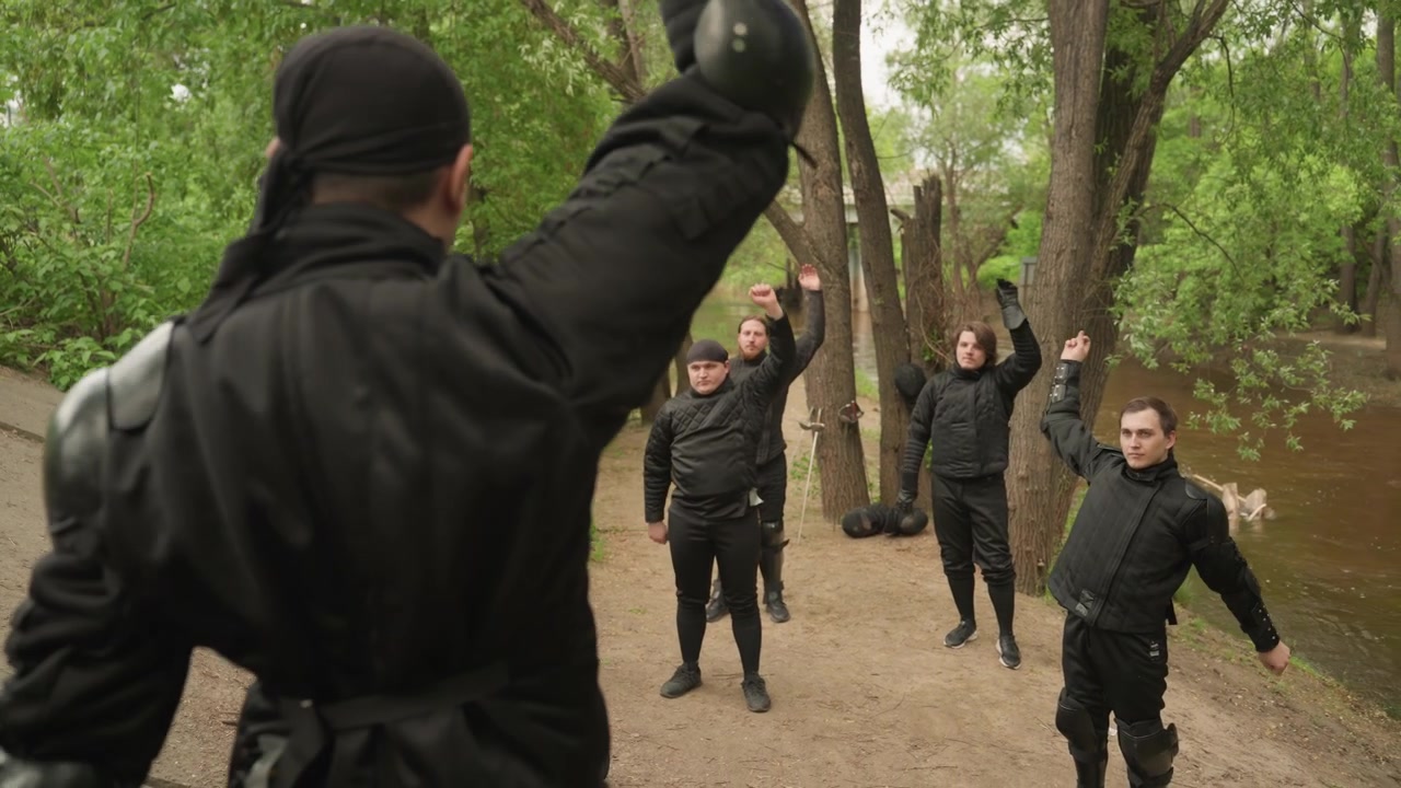 Gruppo di uomini in equipaggiamento protettivo nero che eseguono movimenti sincronizzati all'aperto vicino ad alberi e acqua. Tutti gli uomini indossano tute imbottite con guanti e ginocchiere visibili su alcuni partecipanti. Naturale