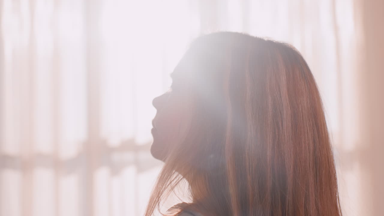 Close up side view of young lady stretching arms behind head with eyes opened in peaceful morning moment, long hair falling naturally, warm light streaming through curtain onto relaxed face