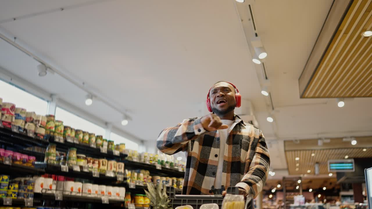 A happy man with Black skin in a plaid shirt with a beard and in red wireless headphones dances and rolls a cart among the counters in a modern grocery store