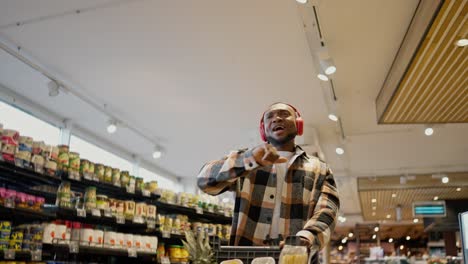 A-happy-man-with-Black-skin-in-a-plaid-shirt-with-a-beard-and-in-red-wireless-headphones-dances-and-rolls-a-cart-among-the-counters-in-a-modern-grocery-store