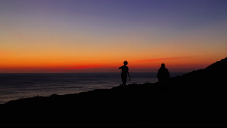 silhouettes of adult walk along shoreline at sunset, wide