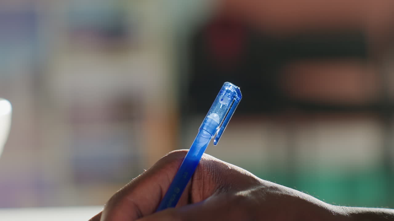 Close up of dark skinned hand holding blue pen while writing in well lit indoor setting, soft background blur emphasizing subject detail, capturing moment of concentration