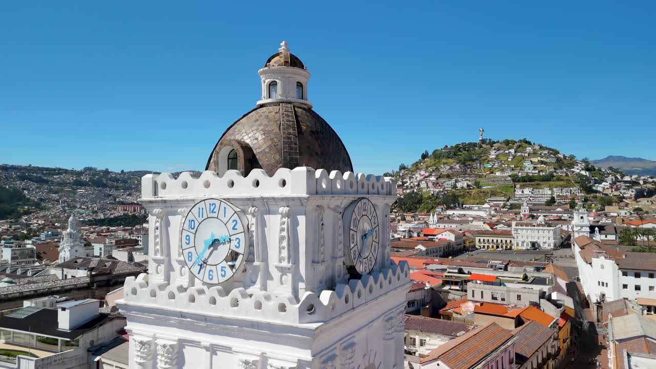 Opportune riprese aeree in parallasse della torre dell'orologio quadrata bianca della Basilica de La Merced con numeri blu e cupola a tegole marroni sullo sfondo del paesaggio urbano di Quito.