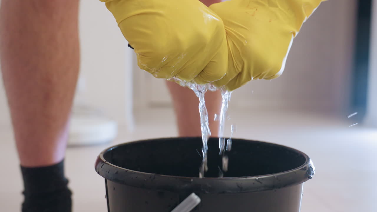Close up of person wearing yellow gloves squeezing wet rag above black bucket with water dripping down, droplets splashing around, cleaning concept