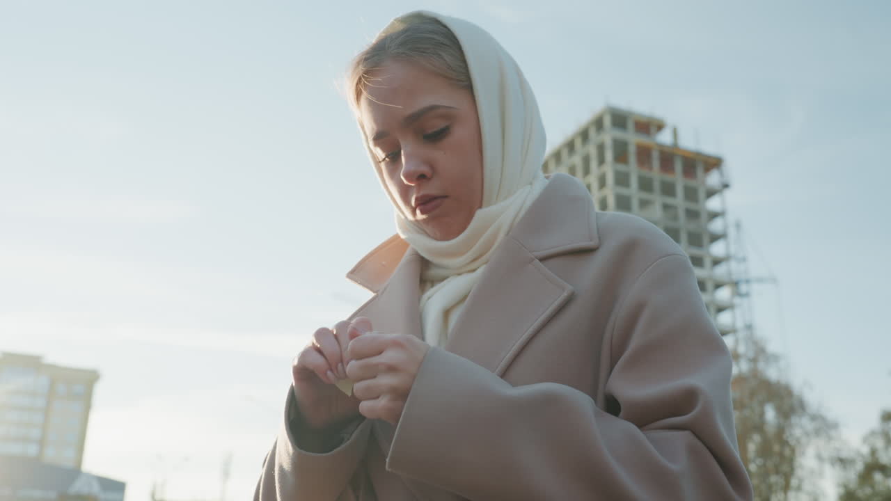 Молодая женщина на улице поправляет шарф, утренняя городская сцена с шерстяным пальто и платком, строительная площадка на заднем плане, мягкий солнечный свет и бледное небо, задумчивое выражение лица с теребящимися руками, медленно.