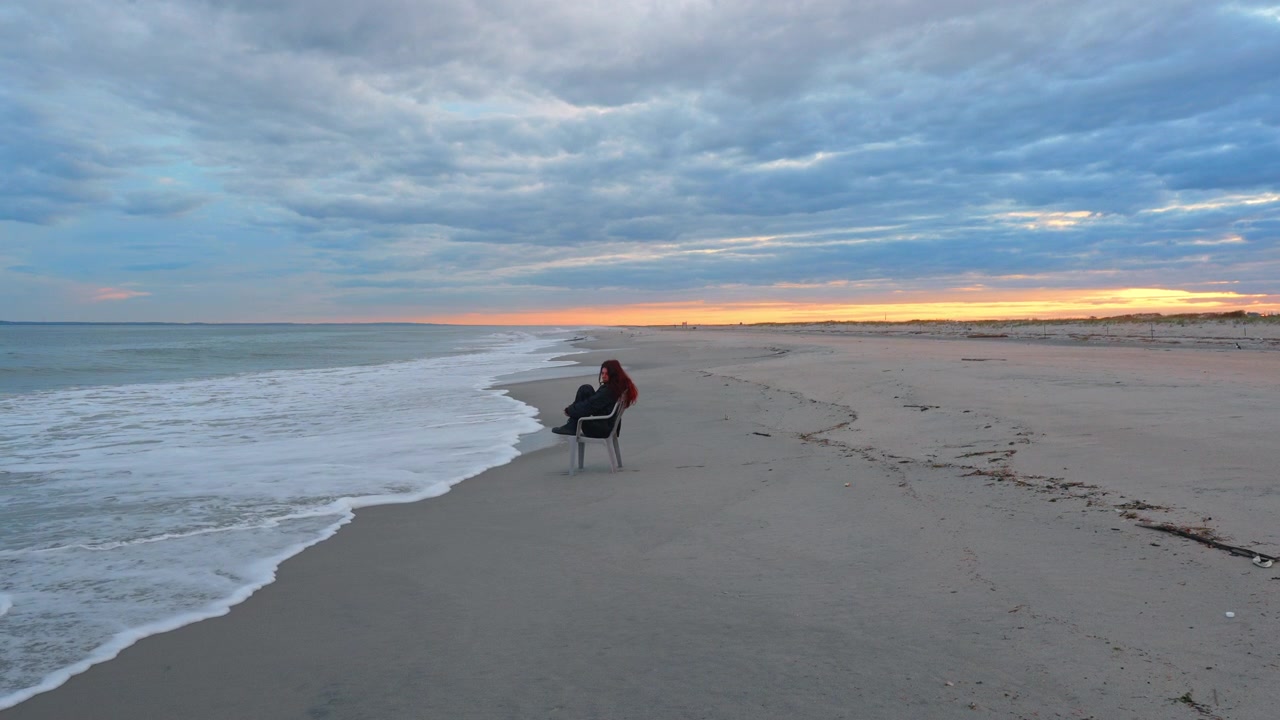 Person sits on a chair by the beach during sunset with waves nearby. A person sits alone on a chair at the beach while the sun sets and waves crash gently.