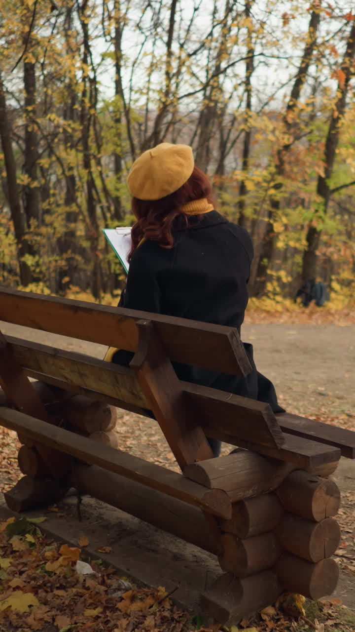 задний вид женщины в желтом берете, рисующей на холсте, сидящей на деревянной скамейке в парке, окруженной осенними листьями деревьев и рассеянной листьями на земле