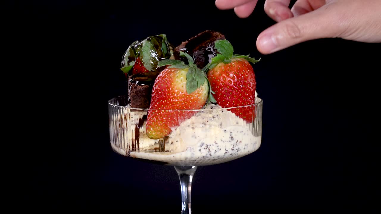 A hand places a large strawberry onto a glass dessert dish filled with vanilla ice cream, chocolate sauce, brownie pieces, and chocolate chips against a black background under studio lighting