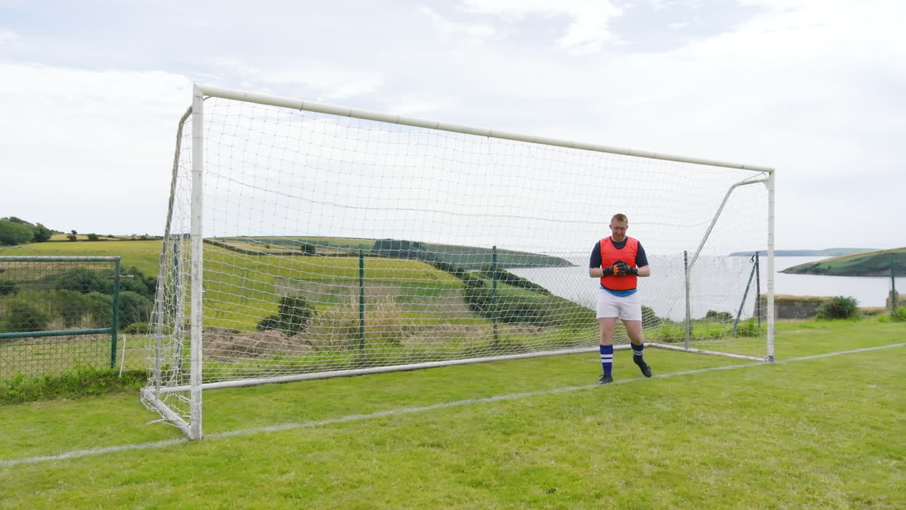 Male goalkeeper wearing orange vest, jumping and defending shoots at goal one after another on pitch