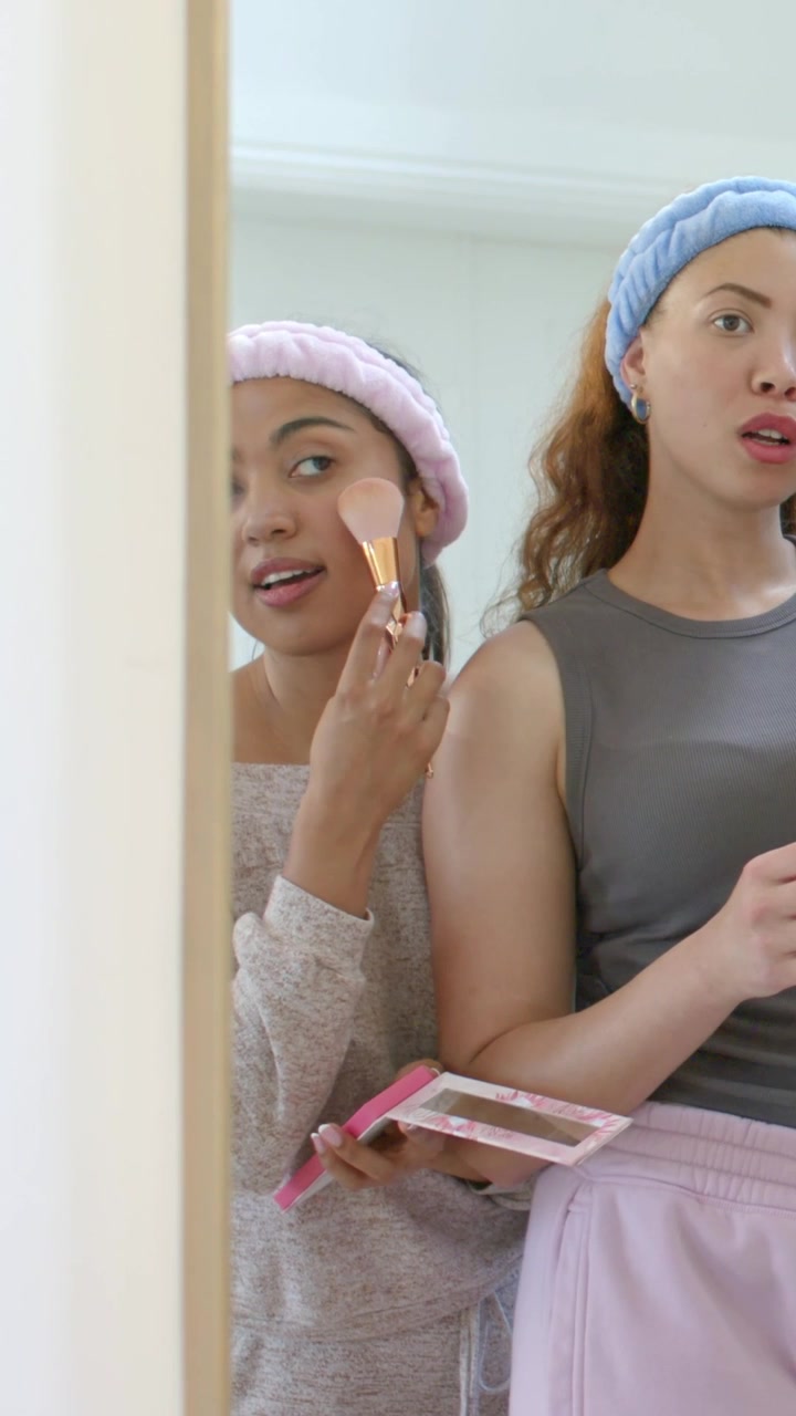 Video verticale: Inizio della routine, due donne che applicano fard e lucidalabbra alla toeletta, indossando fasce per capelli.