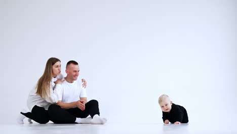 parents sit on a little distance from their baby. mom and dad smile and call their child to get to them.