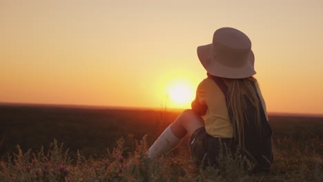 una chica con sombrero admira la puesta de sol en un lugar pintoresco, se sienta en una colina con la barbilla en las manos b