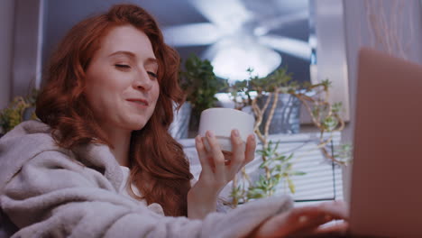 a young caucasian woman is working from home, cozy with a blanket, sipping coffee or tea, happy