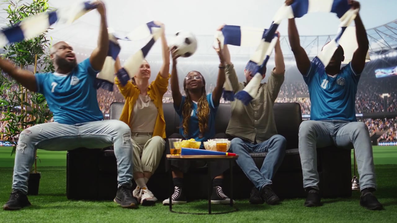 Aficionados al fútbol en el sofá de casa con bufandas del equipo viendo un partido inmersivo.