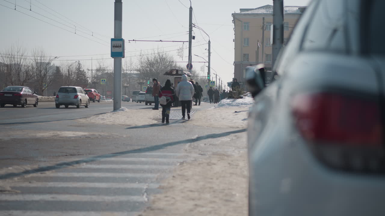 winter city street shows cars parked and moving on both sides while people gather at bus stop, snow edges crossing and poles, cold light, distant buildings and wires frame daily commute moment