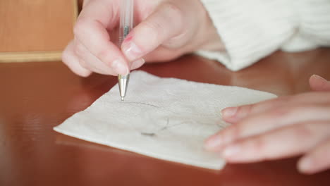 close up of young girl with glasses holding hand-drawn cat mask napkin near mouth while smiling playfully, wearing white sweater, seated against blurred wood and green plant background