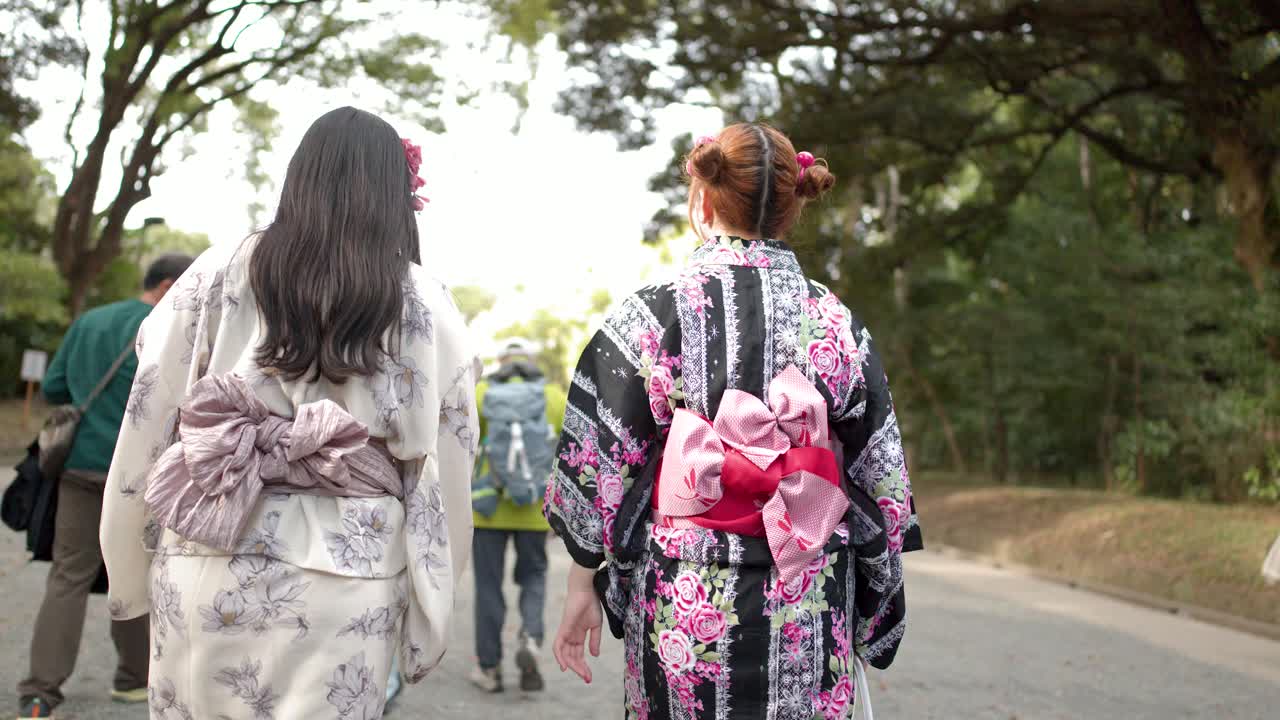 Cảnh nhìn từ phía sau hai người bạn mặc kimono hoa đi bộ qua một công viên nhiều cây xanh ở Tokyo
