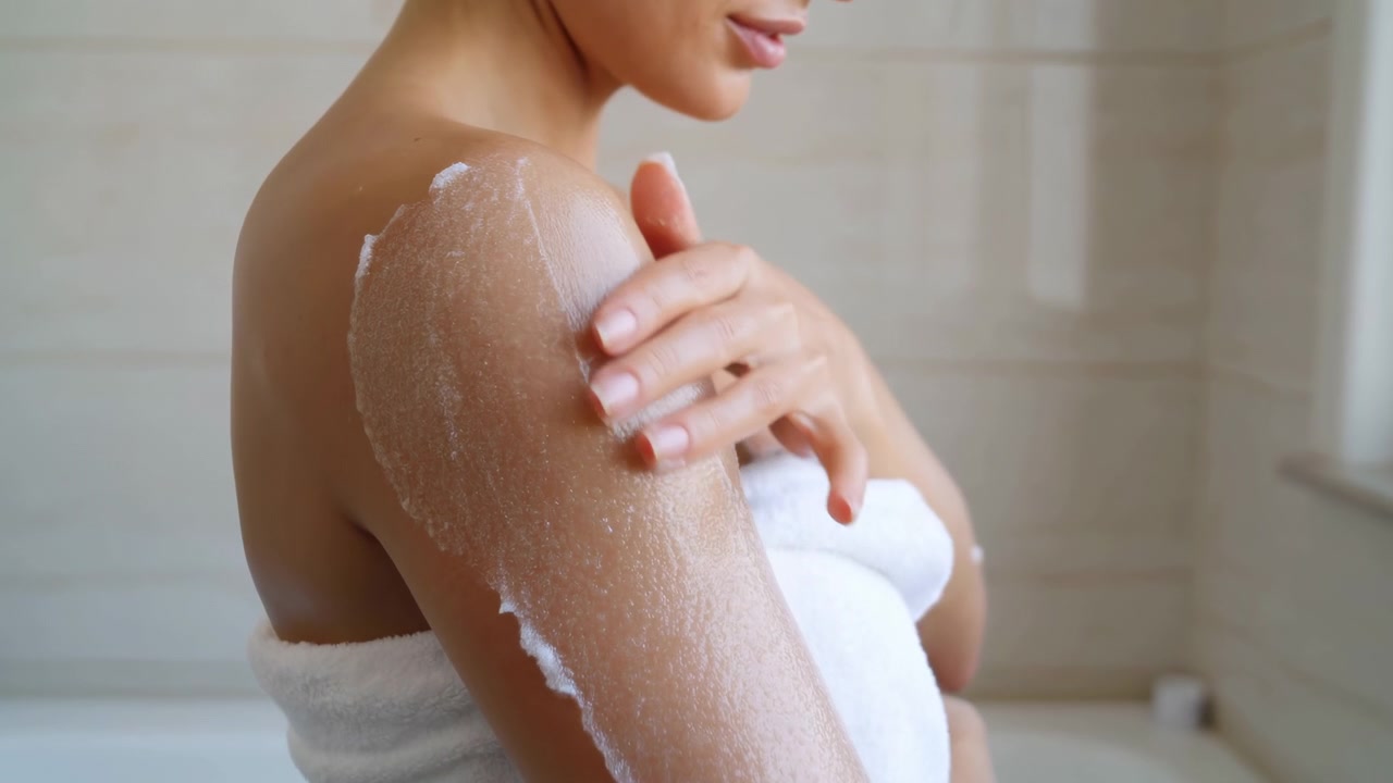 Nærbillede af en kvinde indpakket i et hvidt håndklæde, der påfører eksfolierende scrub på ydre overarm og skulder inde i et neutralt beige flisebadeværelse, hvilket viser blid selvpleje, hudplejerutine og blødt dagslys.