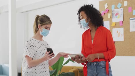 Two-woman-wearing-face-masks-using-smartphone-and-digital-tablet-at-office