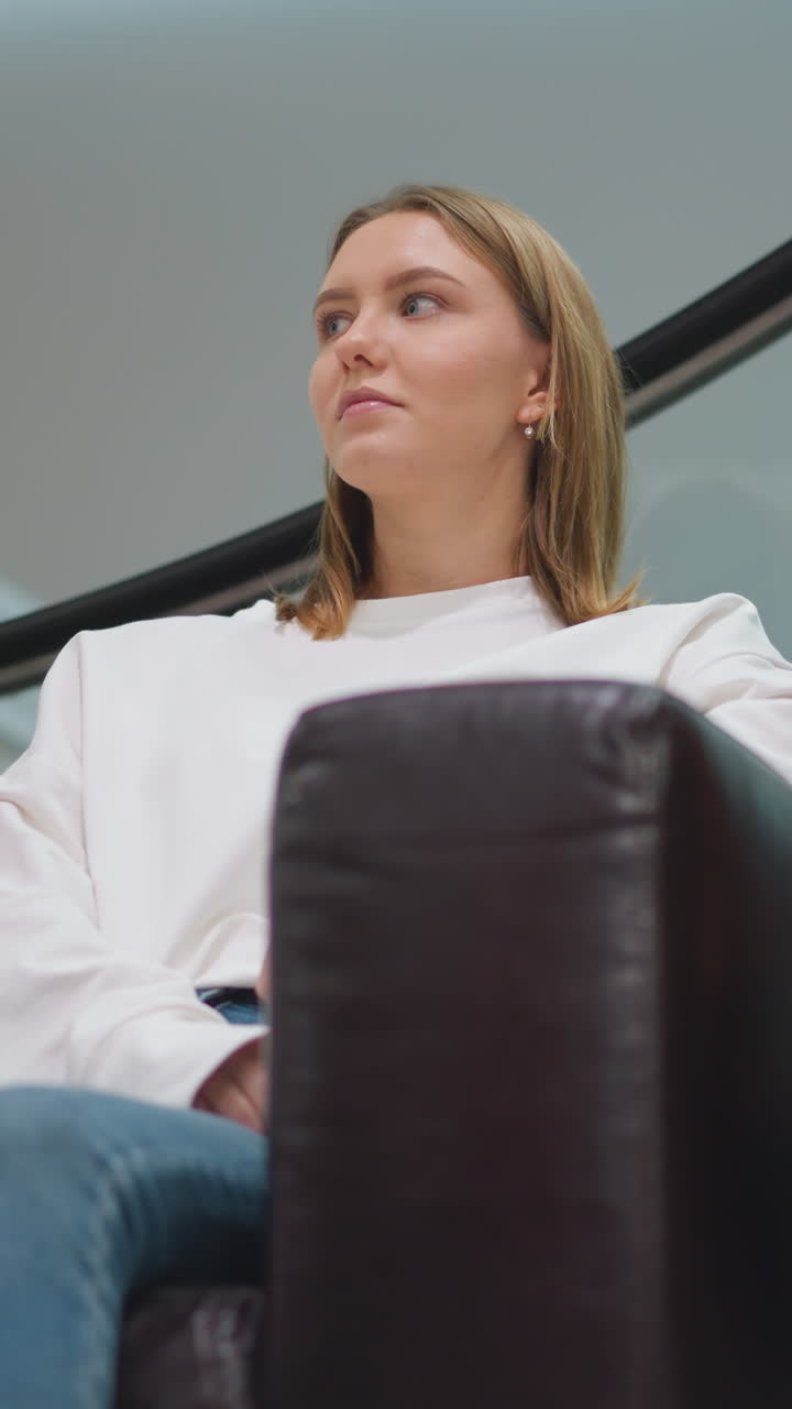 Lady seated on leather chair in modern shopping mall with colorful shopping bags beside her, dressed in casual white top and jeans, with a focused expression, surrounded by stylish fashion displays