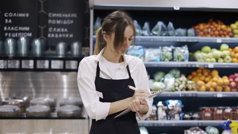 A-woman-worker-in-a-black-apron-and-gloves-carries-out-a-inventory-of-goods-in-a-grocery-store