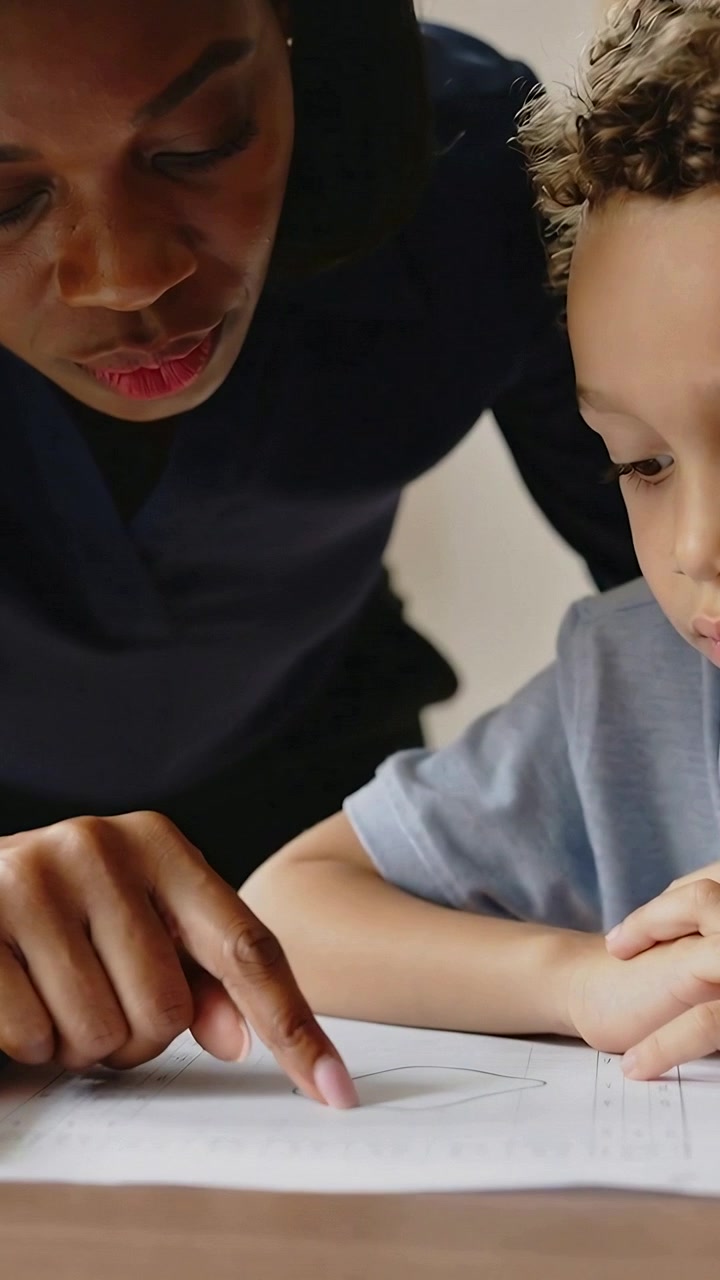 Vertical video: Leaning tutor pointing, tracing heart on sheet at table, guiding child in blue polo
