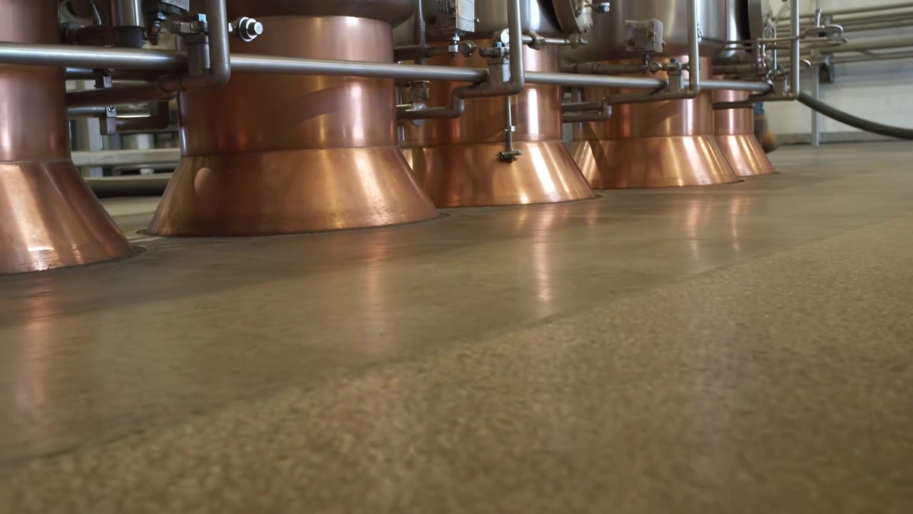 Polished copper equipment and metal pipework slide across frame from floor level while booted legs appear at right, revealing a clean industrial processing room setting.