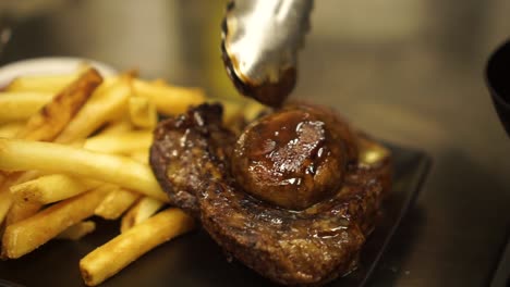 slowmo - close up - finishing the presentation of grilled beef steak, gravy, mushrooms, golden crispy french fries and green salad on a black plate