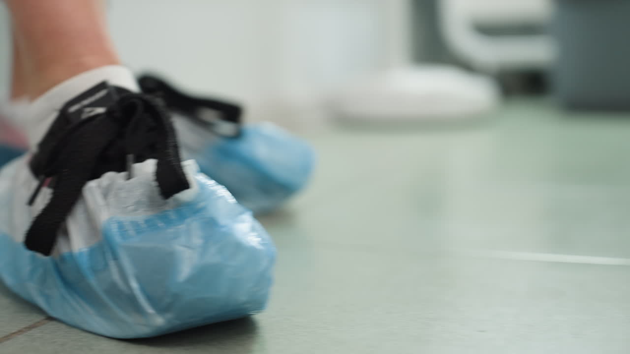 Lady wearing plastic shoe cover walks into bright white clinic room with tiled floor and follows treatment provider to cream massage chair for treatment start, highlighting hygiene