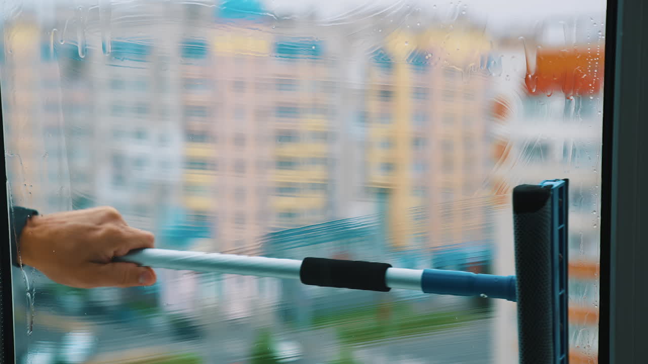 Cleaning dirty window with solution. Worker using household brush to wash window with special liquid inside. Blurred multi-storey building backdrop.