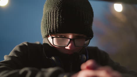 close-up of a young boy wearing glasses and a black jacket, with his head bowed, and a headset around his neck, his hands are clasped and blurred in the foreground