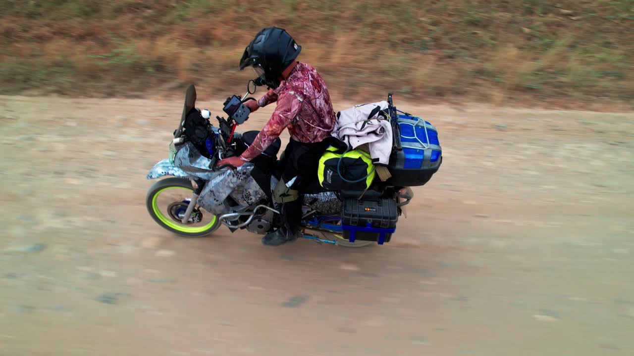 Man on a heavily loaded adventure motorcycle rides on a dirt road, with dry, sparse vegetation in the background