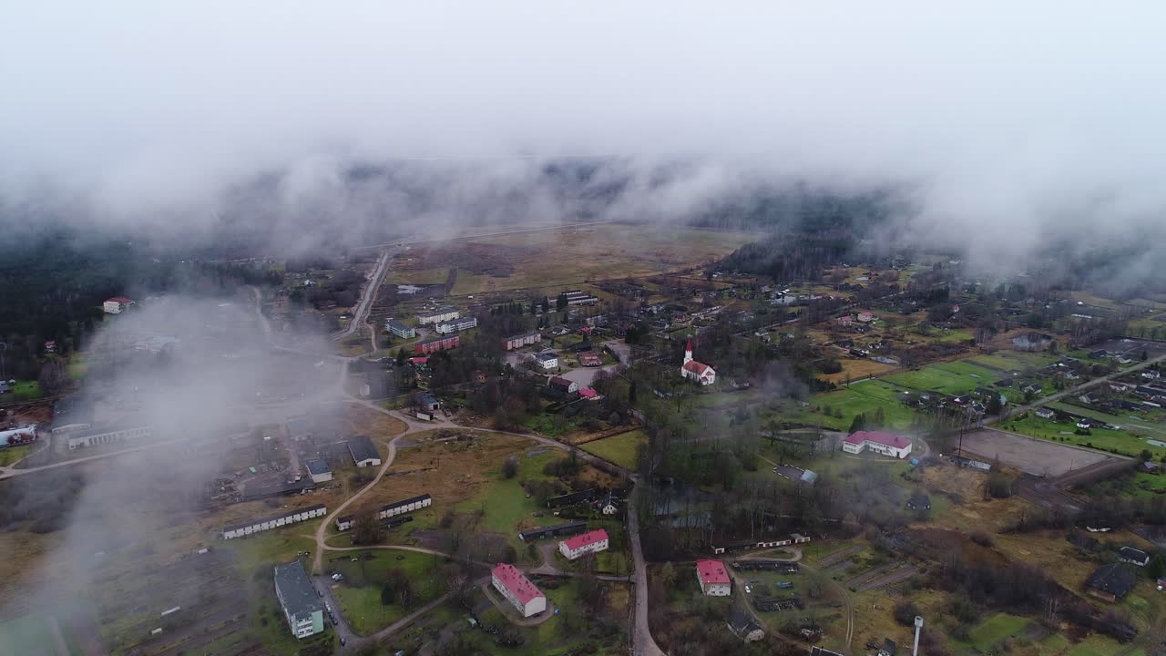 Aerial flyover of fog-covered Rucava village under dark clouds and misty air