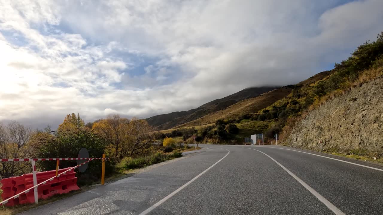 A car travels along a winding farm road through autumn hills and fields in Queenstown, New Zealand, under dramatic, partly cloudy sunlight