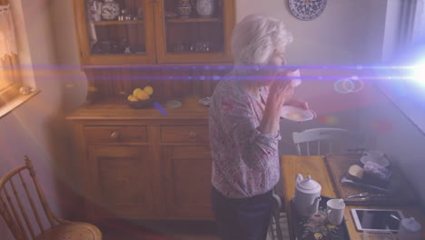 animation of light moving over senior caucasian woman in kitchen looking out of window drinking tea