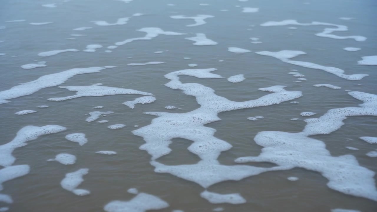 Showing foamy shallow surf shifting across wet sand as small waves and backwash move foam apart