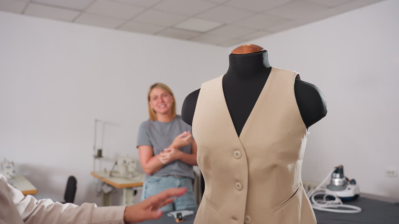 Clothing designer places hands on beige sleeveless jacket worn by mannequin while smiling and laughing joyfully with partner in shared moment of celebration after completing fashion project together