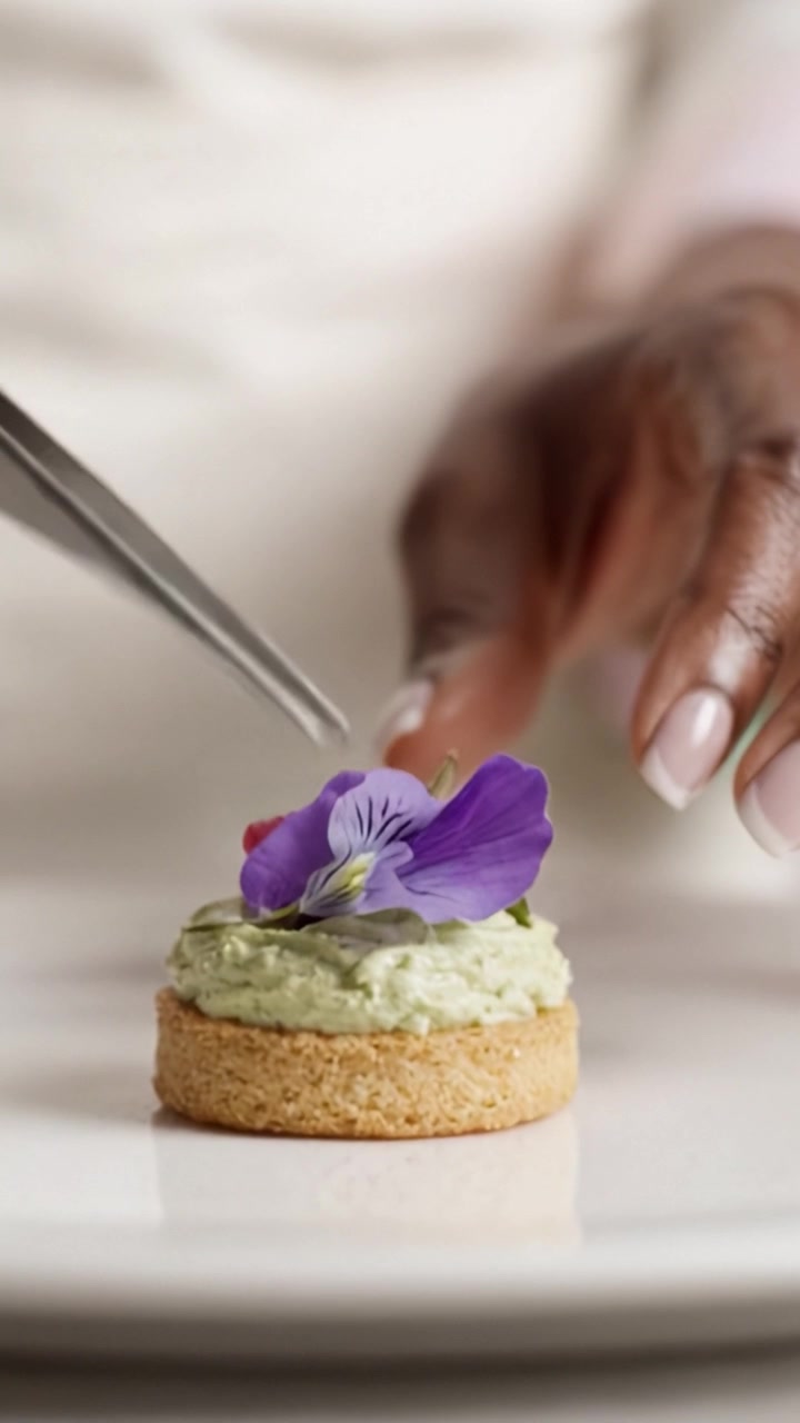 Kulinarisk kunst: En delikat hånd tilbereder en smuk dessert med spiselige blomster og cremet glasur, hvilket viser færdighed og passion for gastronomi.