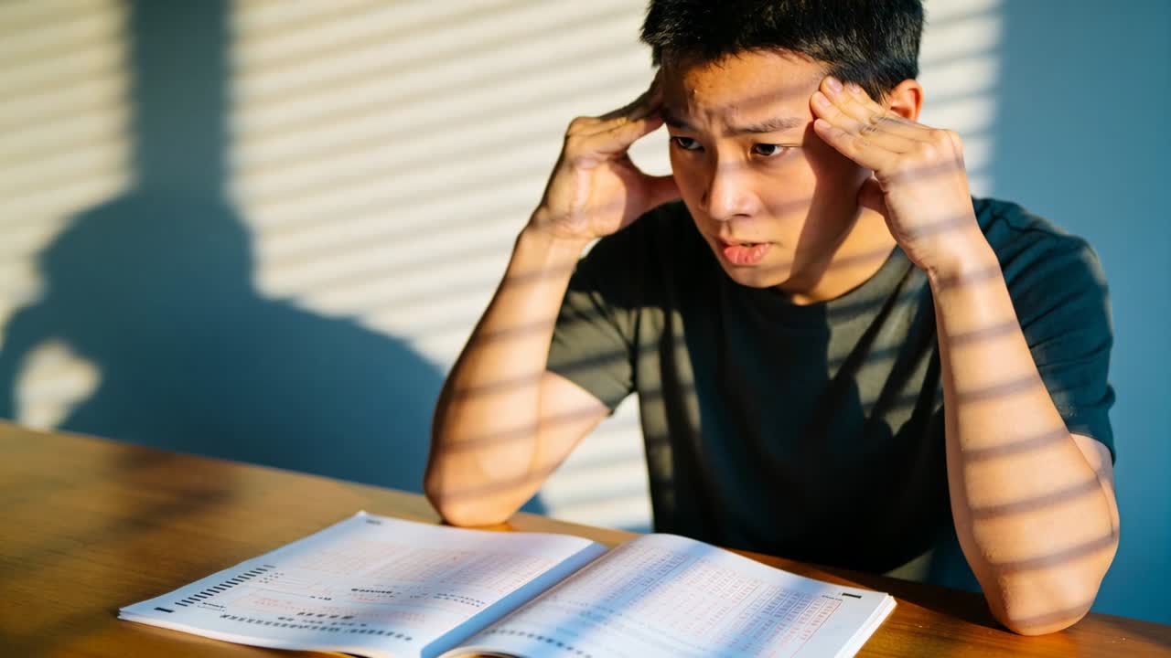 Facing hard workbook problems, Asian teen rubbing temples, squinting at wooden table by blinds