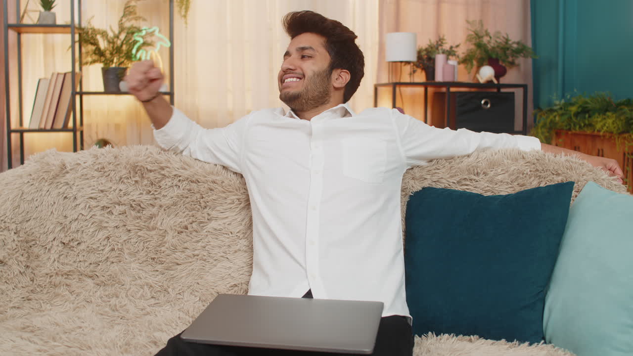 Indian man smiling with satisfaction closing laptop celebrating success feeling accomplished on sofa