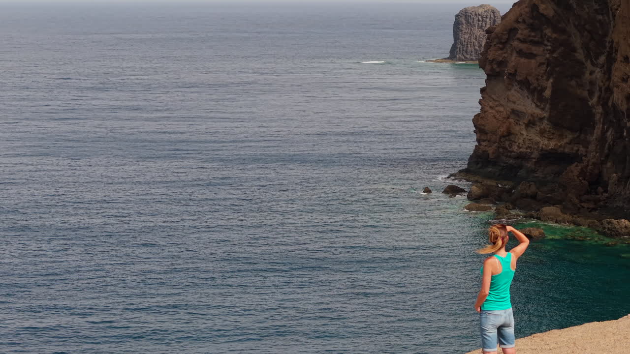 Female tourist standing on rocky cliff edge, overlooking expansive ocean panorama in Gran Canaria, Spain, capturing serene coastal landscape