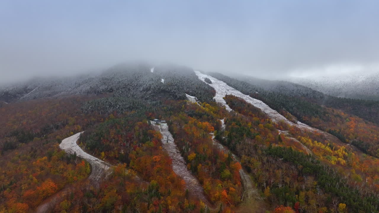 Tops of the mountains covered with thick mist. Drone footage descending above the mountains covered with woods colored in red, orange and yellow. Meadows in the forests are sprinkled with first snow.
