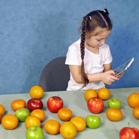 little girl holding tablet in the kitchen. sweet fruits laying on the table. cute child playing games on gadget near the table with fresh apples and oranges.
