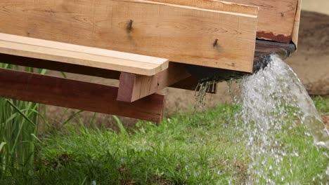 a wooden trough directs a stream of water onto lush green grass, creating a serene outdoor scene.