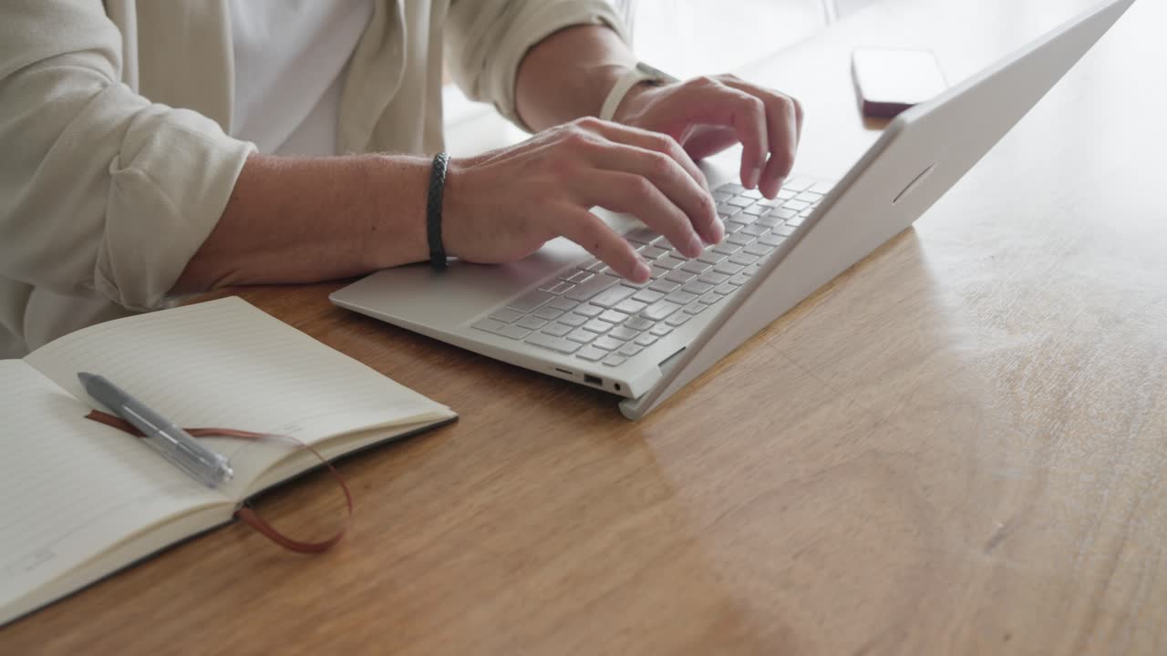 Uomo adulto appoggia le dita sulla tastiera del portatile, digita, inclina lo schermo mentre lavora al tavolo con degli appunti